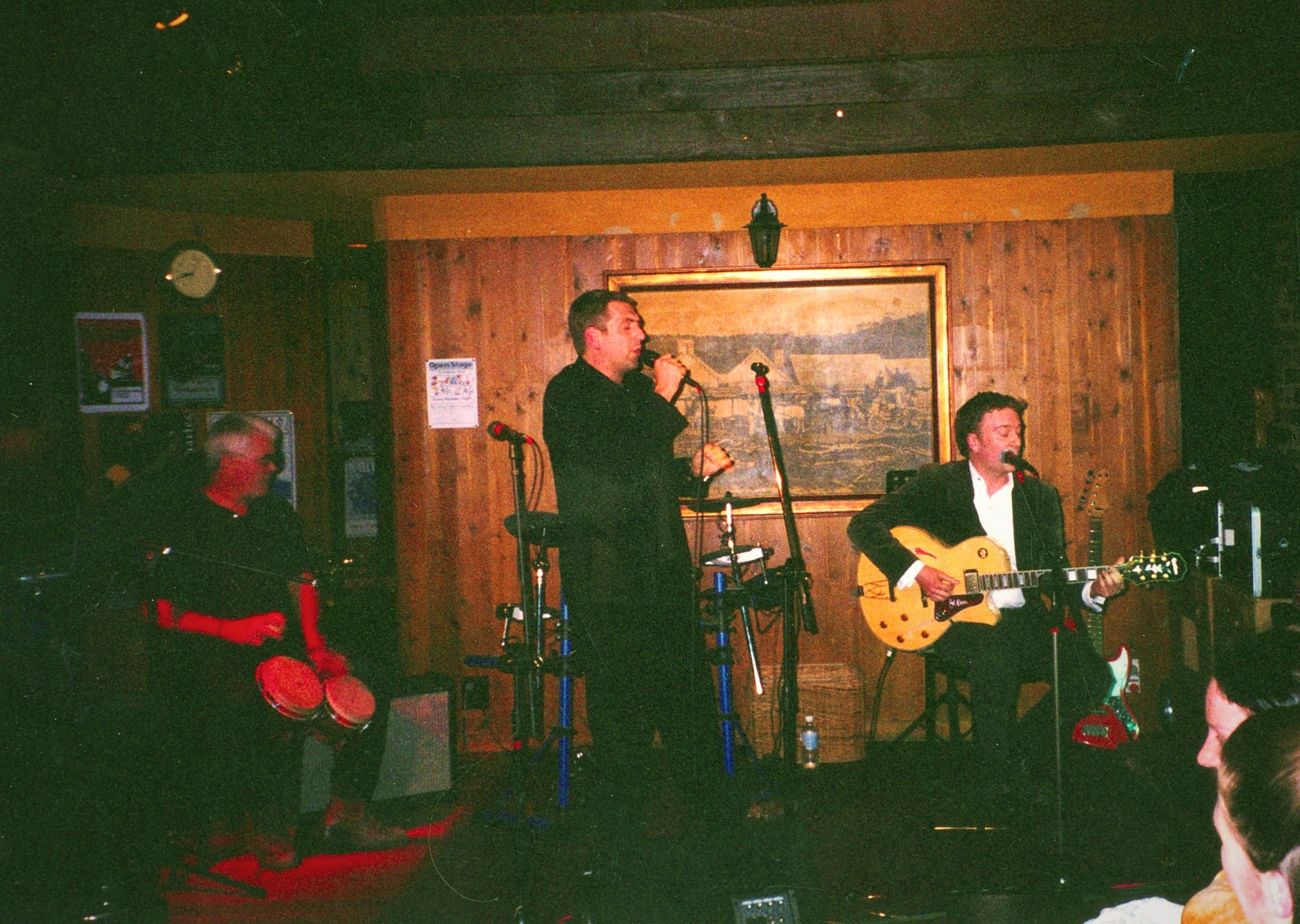 Frankie Velvet at the microphone in a wood-panelled room, a percussionist to his left and a guitarist with a gold-top archtop to his right.
