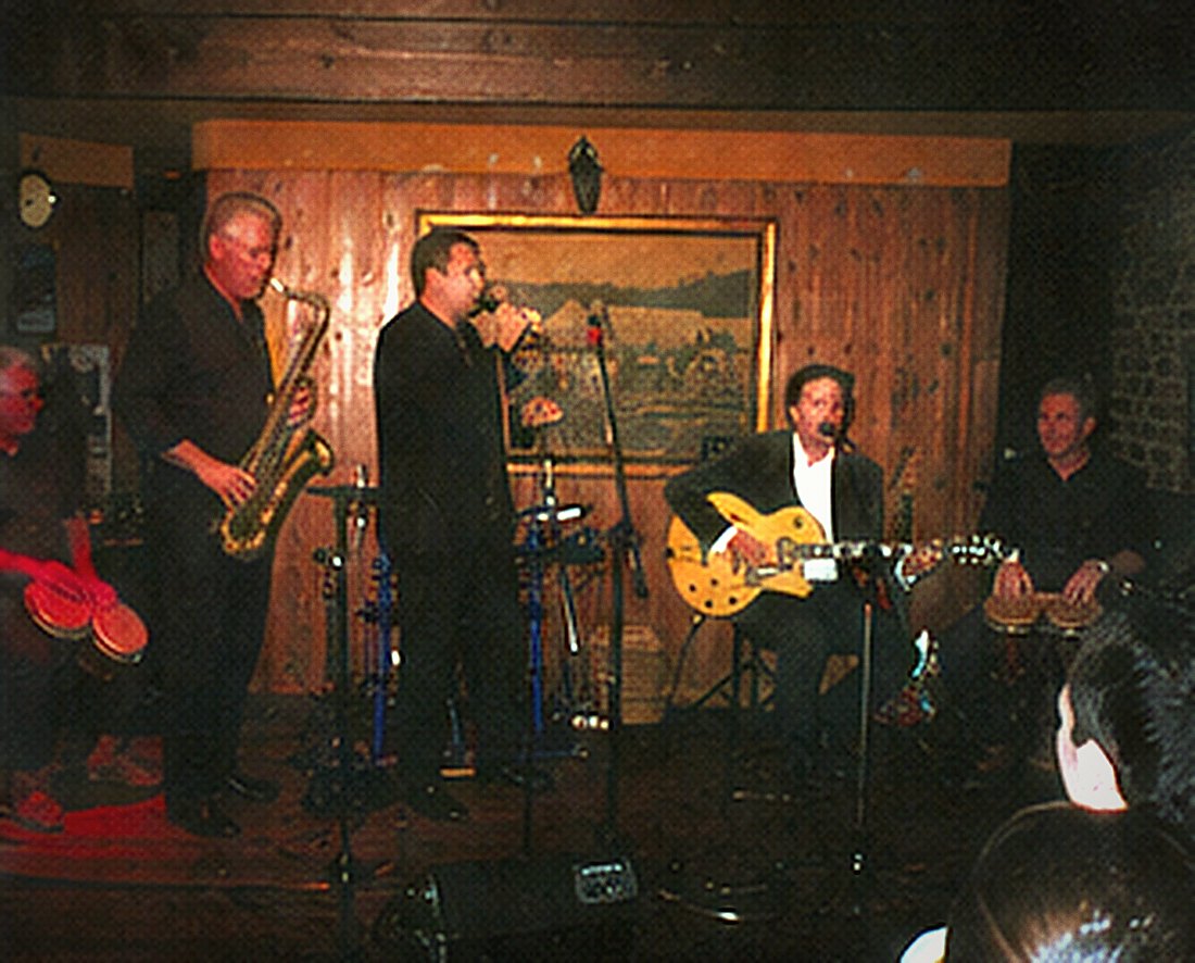 Five musicians on a small stage: percussion, tenor saxophone, singer, guitar and a second percussionist.