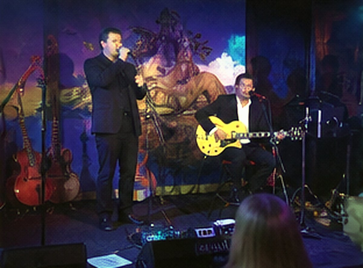 A singer and guitarist on a blue-lit stage before a painted backdrop.