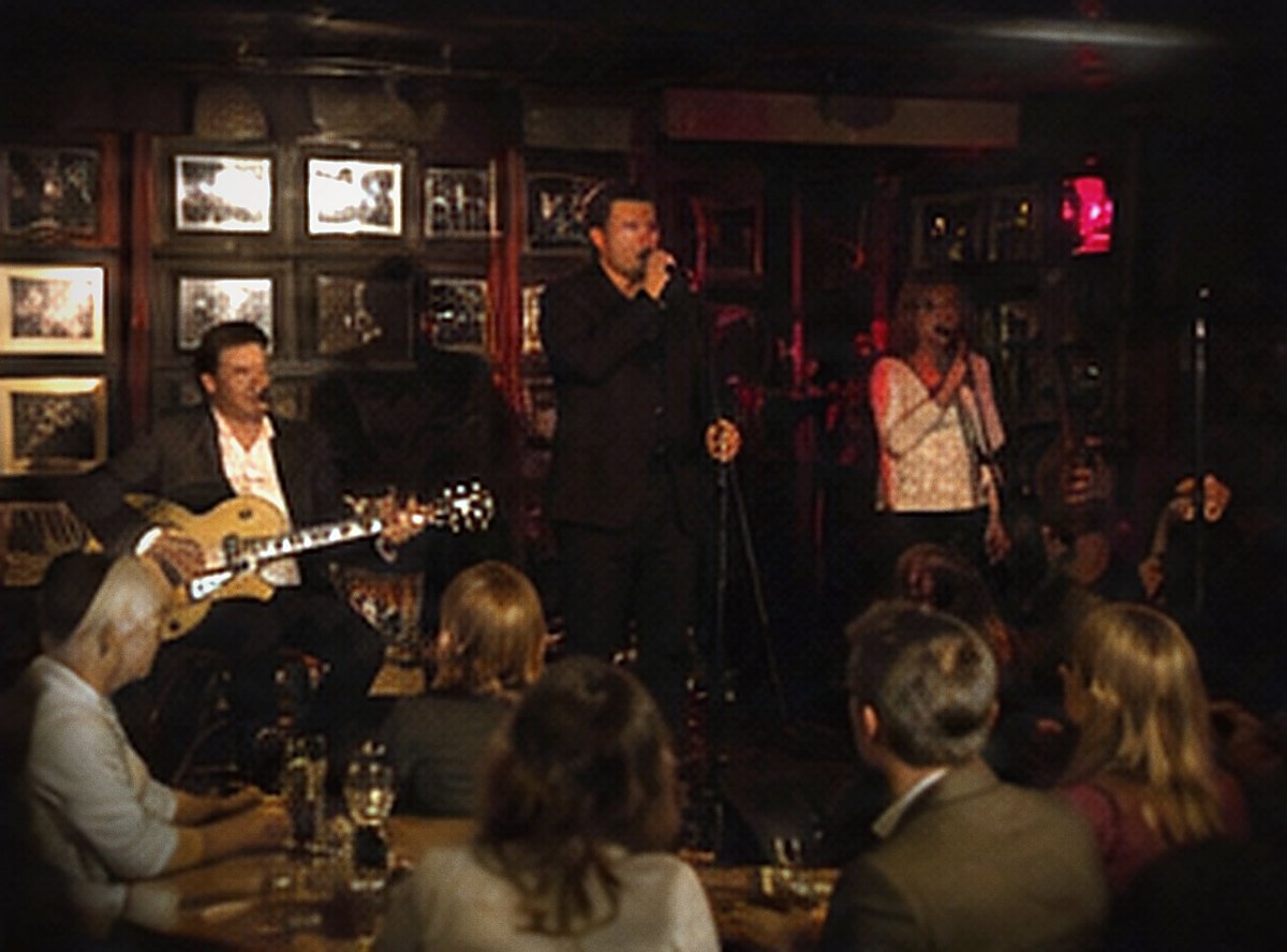 A dark club room, band at the back, tables of seated drinkers in the foreground.