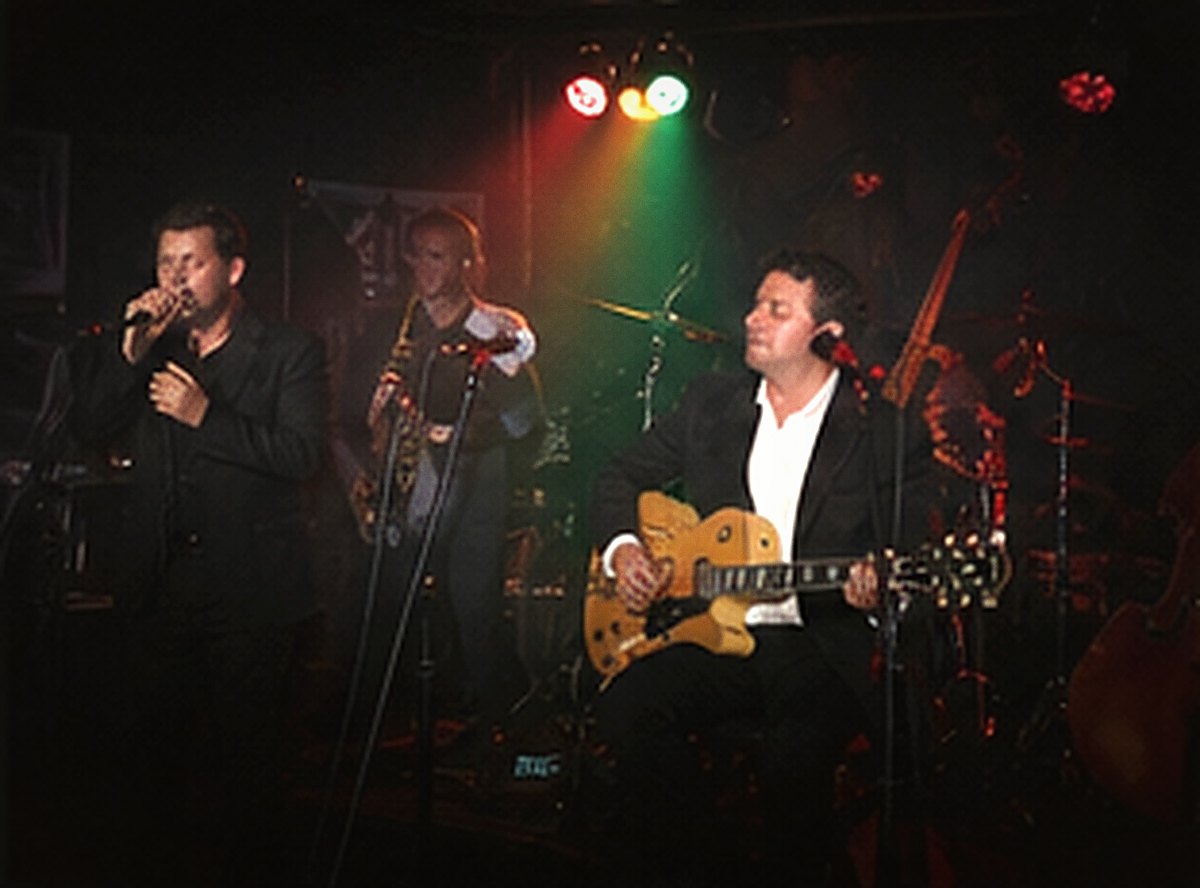 A band lit by green and red stage lights: singer, saxophone and guitarist.