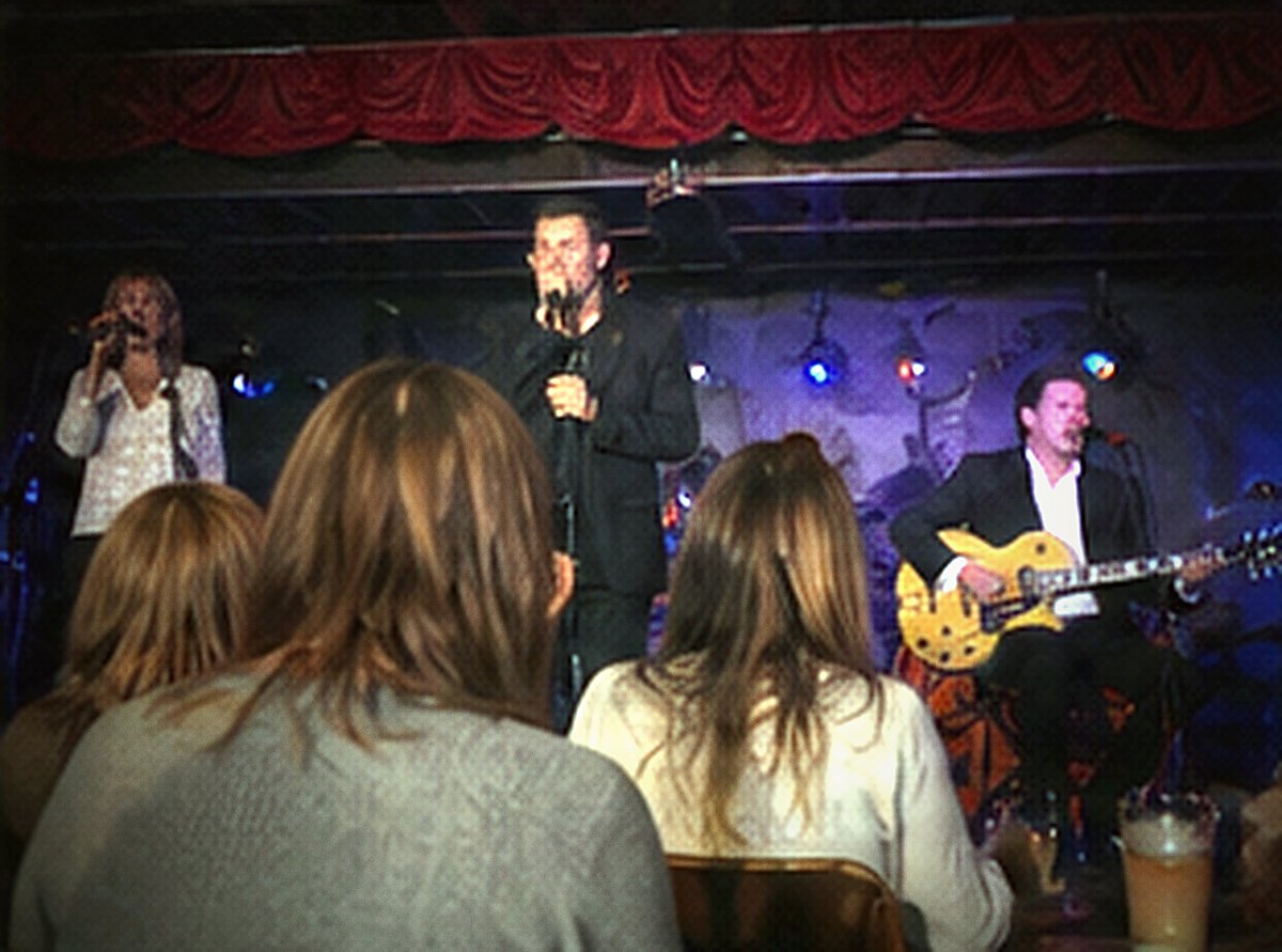 An audience seen from behind, heads close to camera, the band small and bright beyond them.