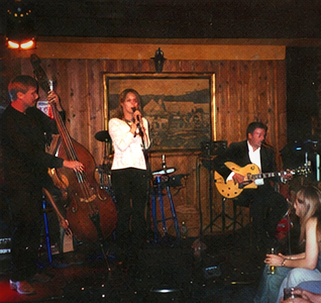A double bass player at the left of a small stage with a singer and guitarist beside him.