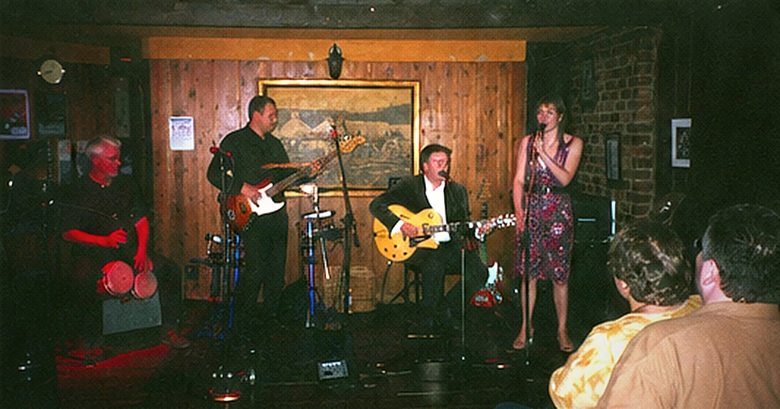 A vocalist in a patterned dress at a microphone, with a bass player and guitarist behind.