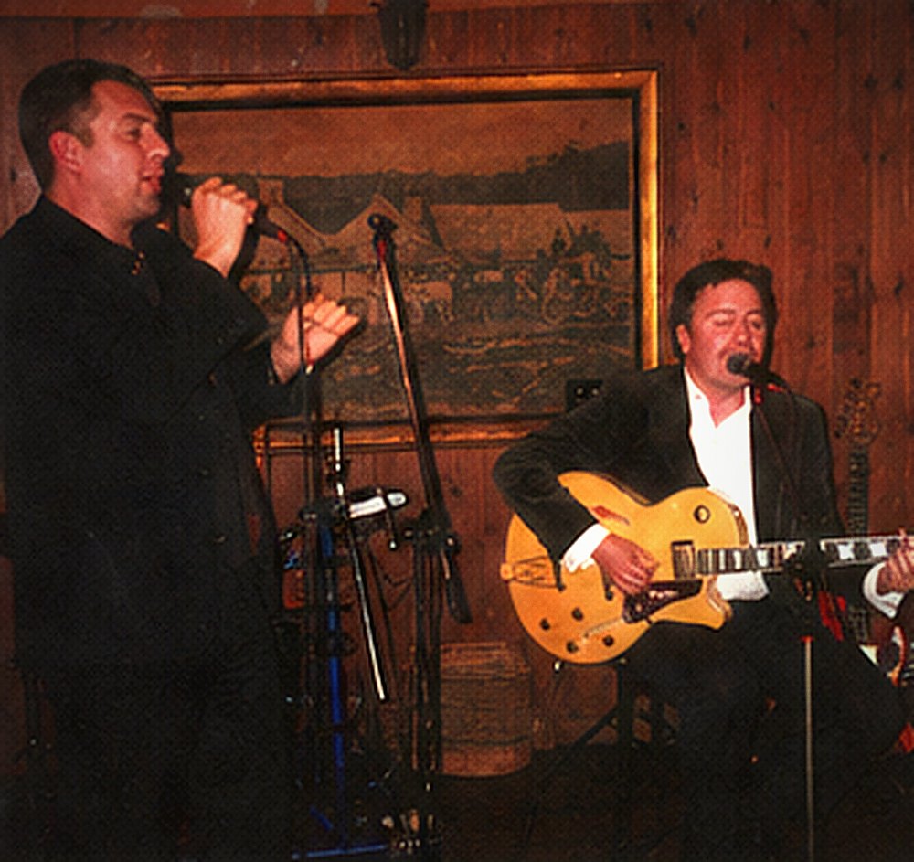 Frankie Velvet singing into a hand-held microphone in profile, the guitarist seated behind him.