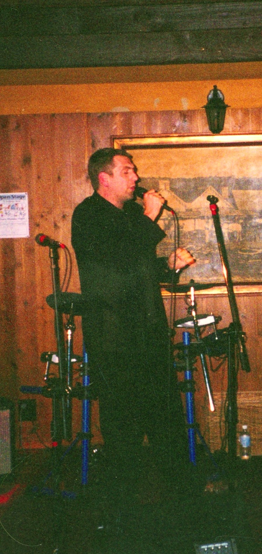 Frankie Velvet in a dark suit singing into a hand-held microphone, full length, against wood panelling.