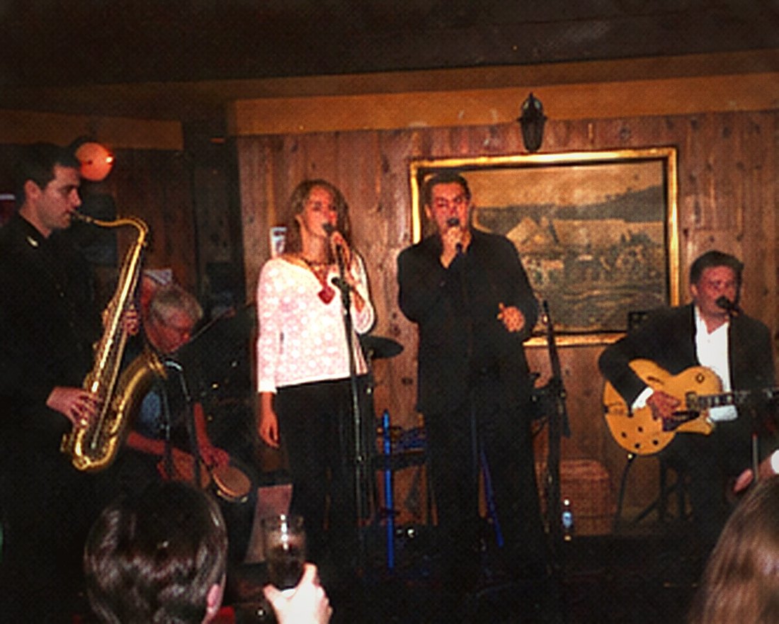 Four performers in a line: saxophone, vocalist, singer and guitarist.