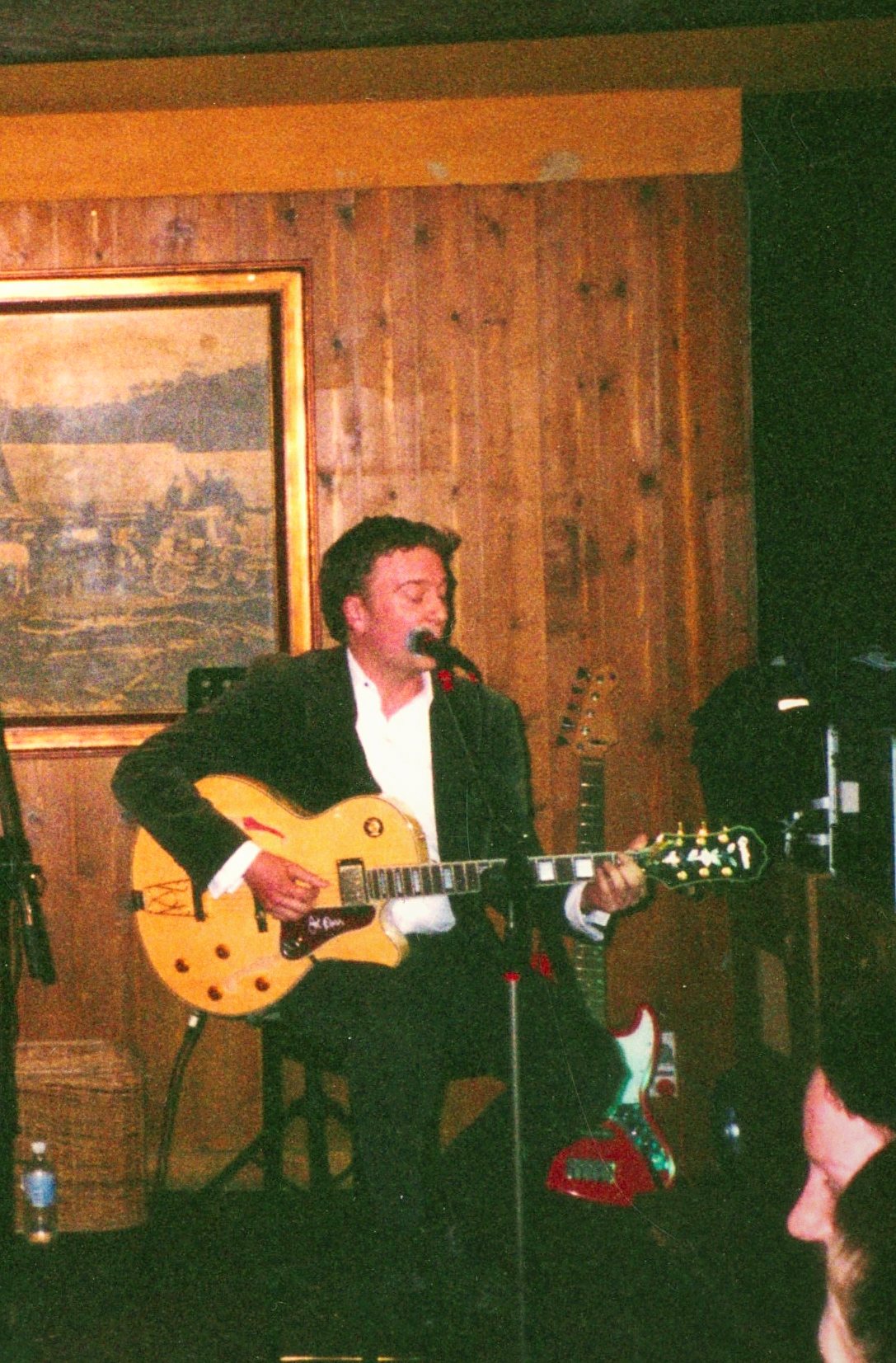 A guitarist in a dark suit and white shirt playing a gold-top archtop guitar under a wall lamp.