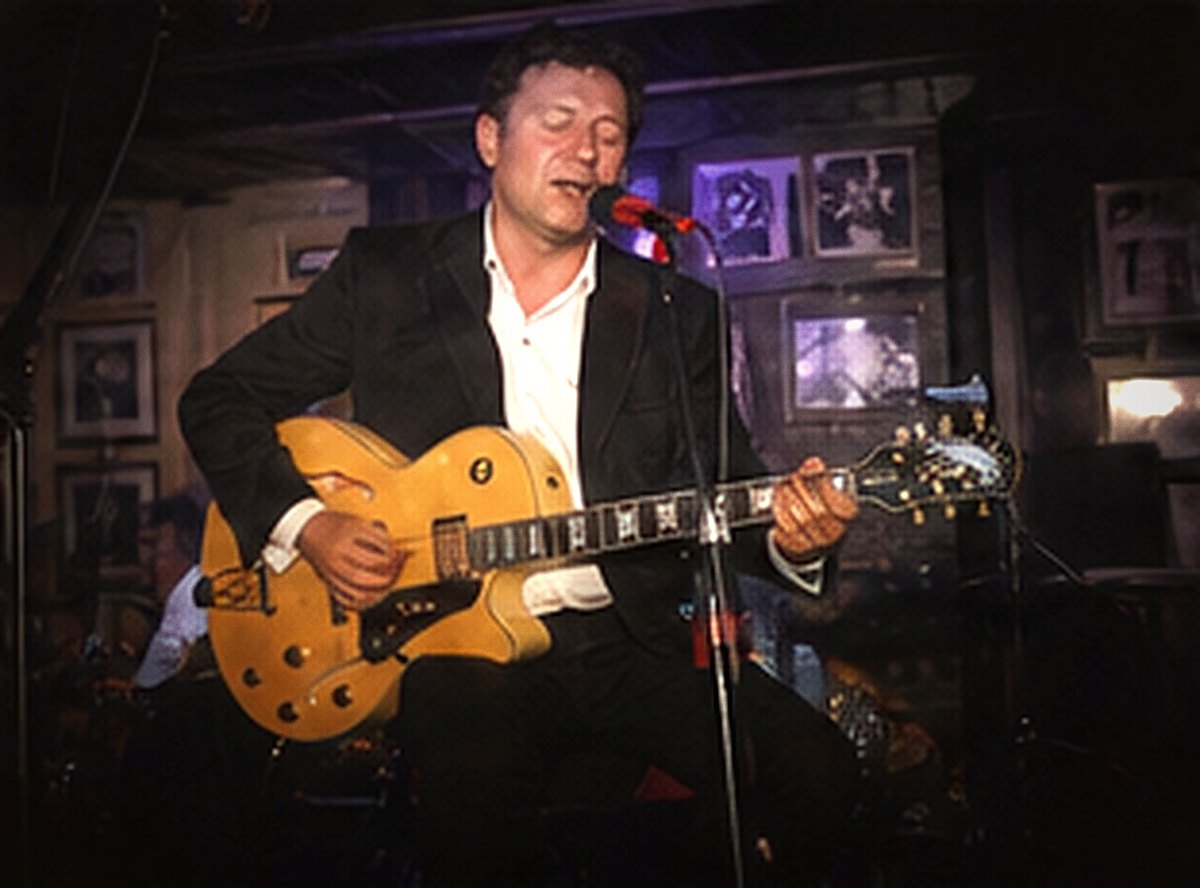 A guitarist in a dark jacket and white shirt, eyes closed, playing a gold-top archtop guitar.