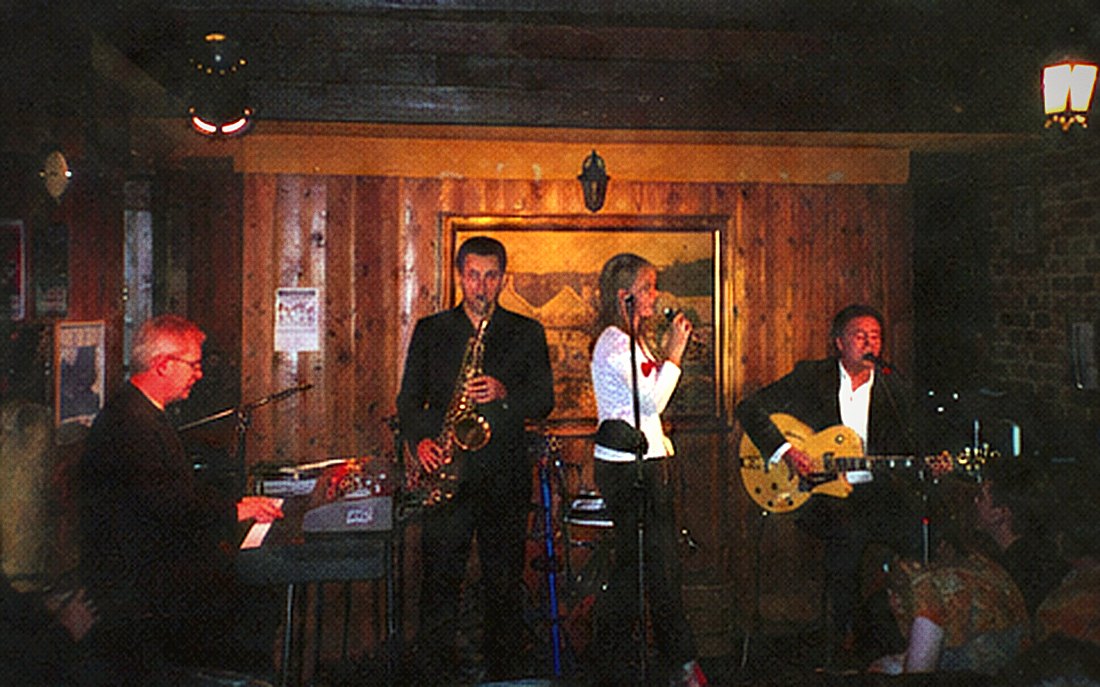 A keyboard player at the left of a small stage with a saxophonist, singer and guitarist.