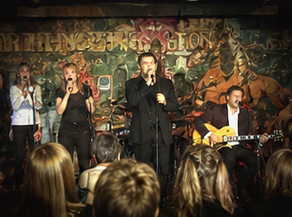 Three vocalists and a singer across the front of a stage before a painted mural.