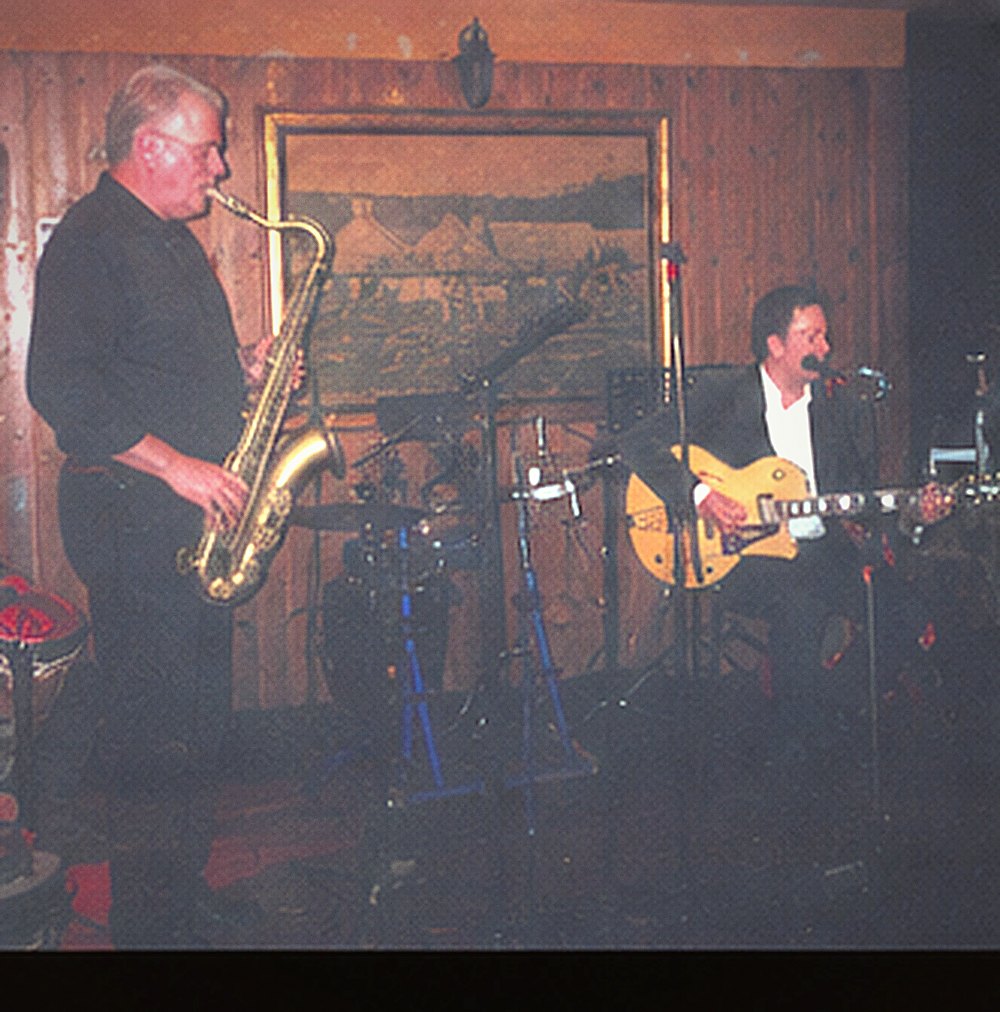 A grey-haired saxophonist in black playing tenor saxophone, lit warmly against wood panelling.