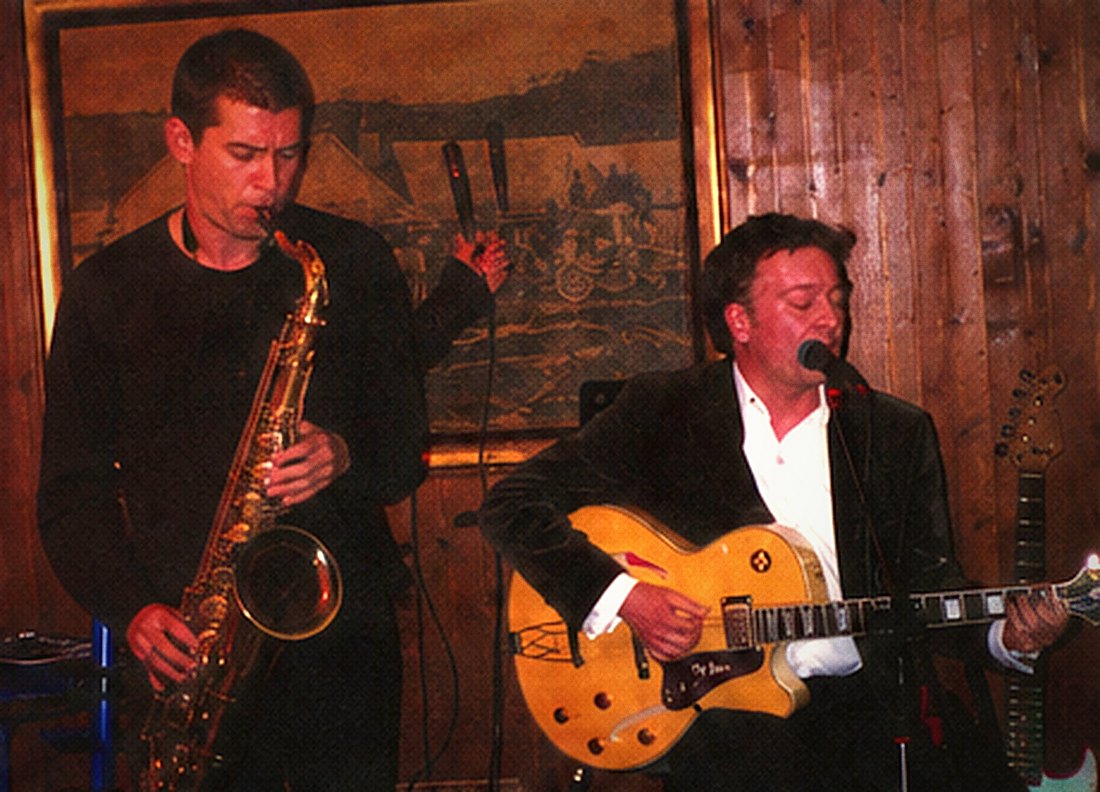 A saxophonist and a guitarist performing side by side under warm stage light.