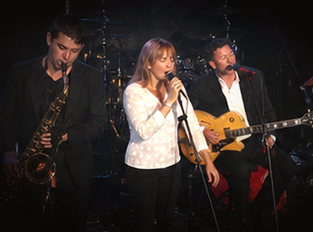 A saxophonist, a vocalist and a guitarist tightly framed together on a small stage.