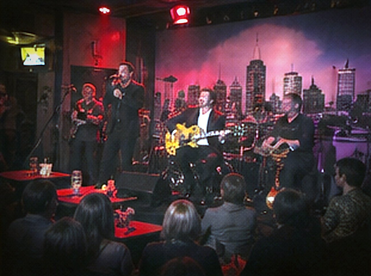A band on a low stage before a painted city skyline with red-lit tables in front.