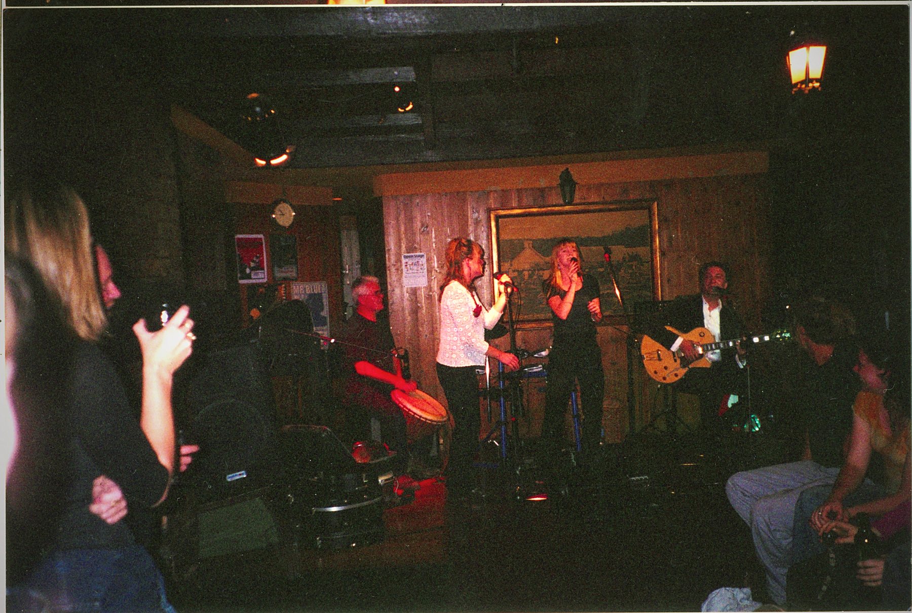 A wide view of a dim panelled bar room with a band performing and patrons watching.