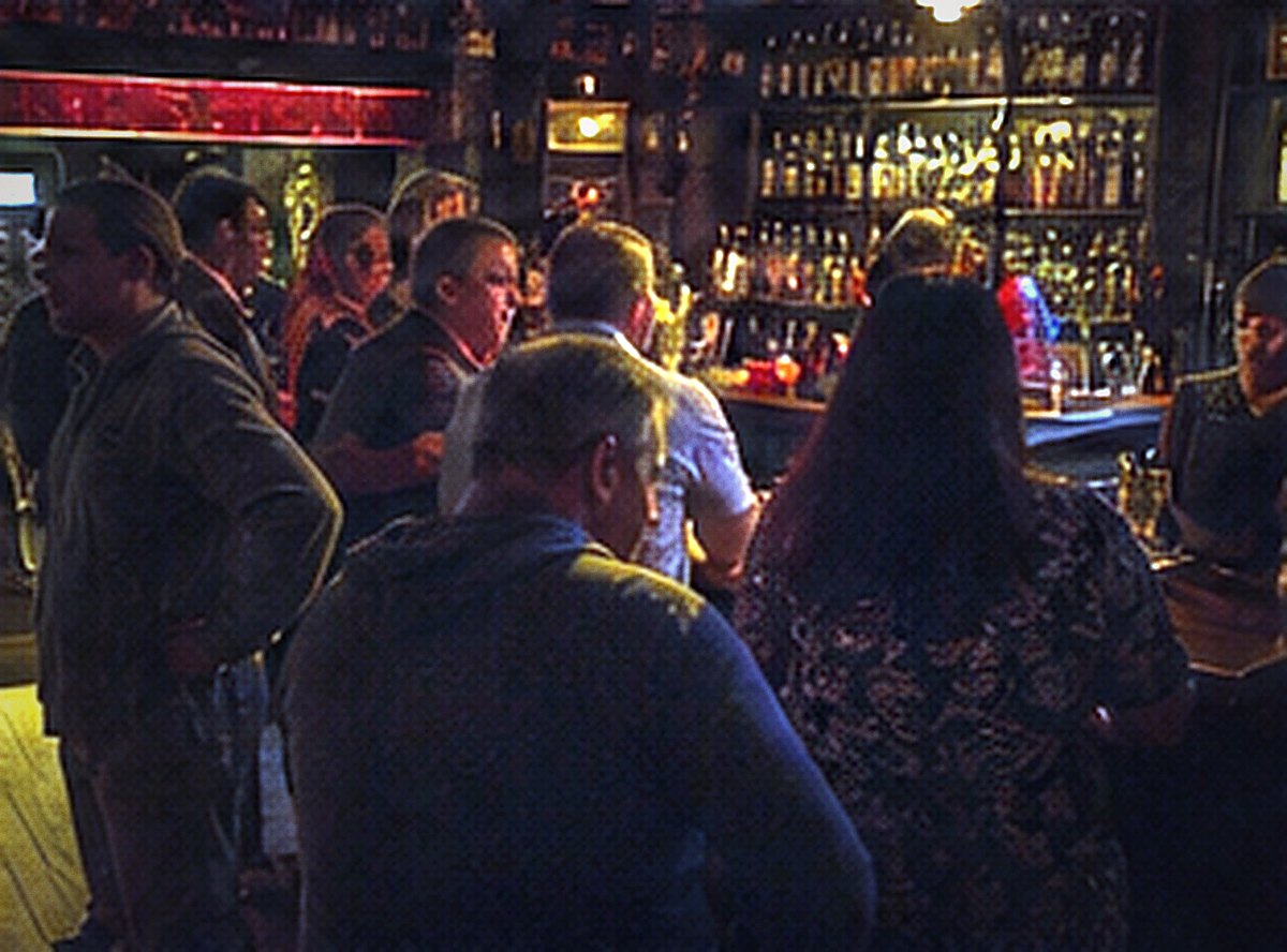 A crowded bar seen over the shoulders of patrons, bottles lit gold behind it.