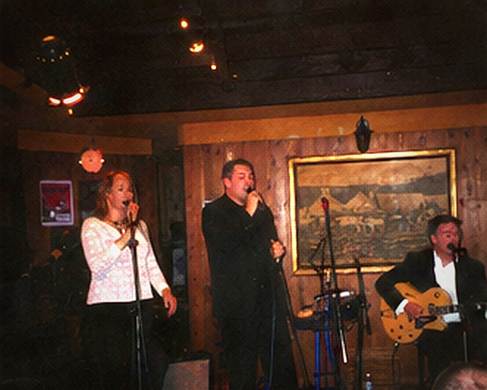 A vocalist and the guitarist singing together at adjacent microphones on a small stage.