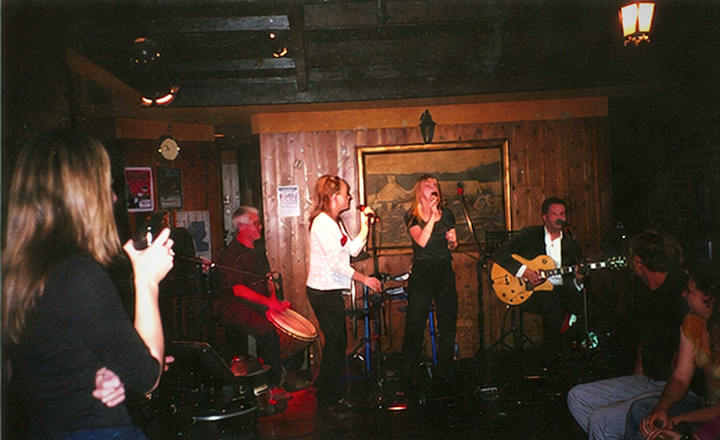 A dim panelled room with a band on a low stage and an audience seated close in the foreground.