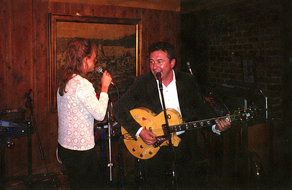 A singer with her back to the camera and a guitarist singing into a microphone opposite her.