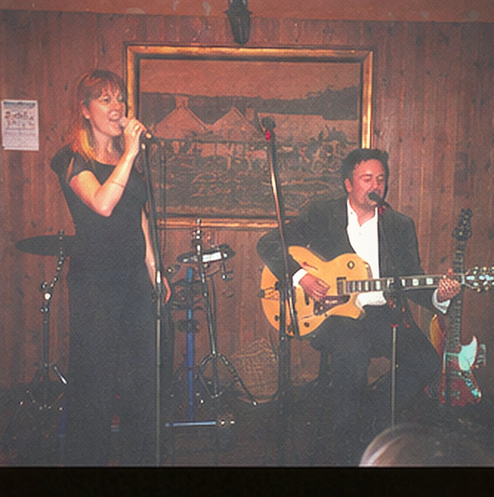 A vocalist in black singing with eyes closed at a microphone, guitarist behind her.