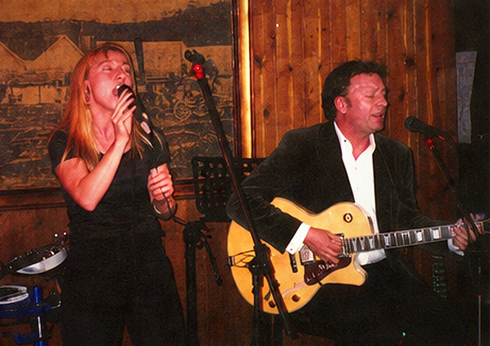 A vocalist singing with head tilted back beside a guitarist.
