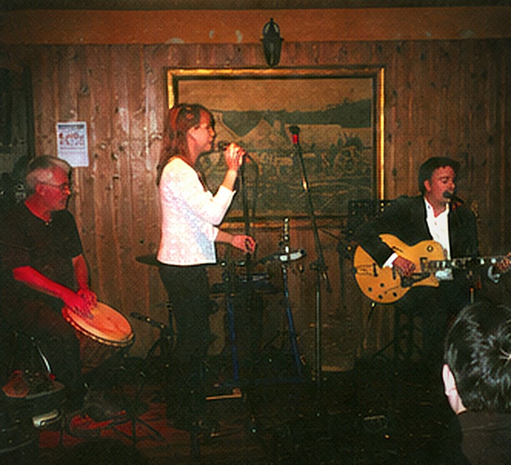 A vocalist singing at the front of a small stage with a percussionist and guitarist behind her.