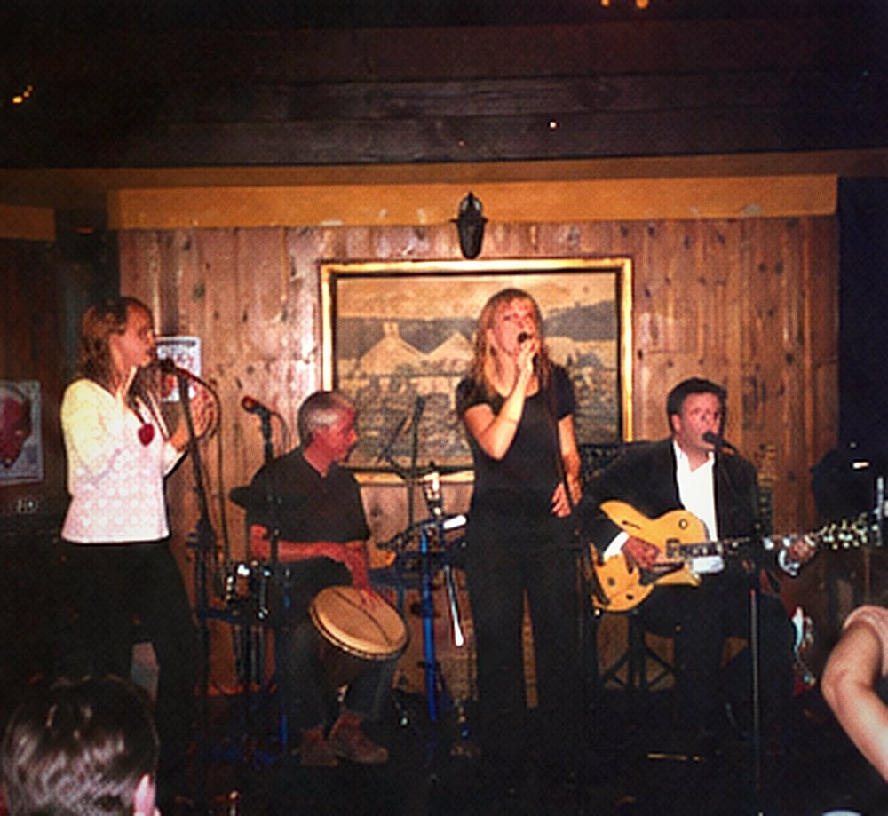 Two women singing at microphones with a percussionist behind them.