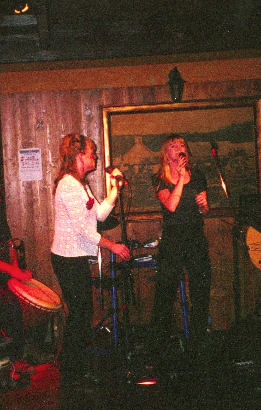 Two women singing into microphones on a small stage, one in a white blouse, one in black.